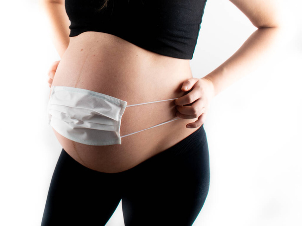 Pregnant woman holding white medicinal face mask on her pregnant belly abdomen. Coronavirus covid19 new normal rules and regulations, isolation and new lifestyle concept - Photo, Image