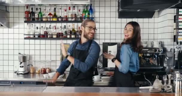 Caucasian young cheerful couple of bartenders in aprons dancing funny and joyfully at counter in cafe. Handsome waiter and beautiful waitress having fun happily in bar at work. Man and woman coworkers - Footage, Video