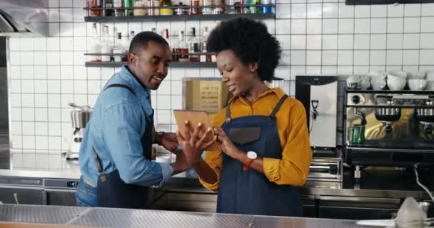 Önlüklü güzel Afro-Amerikalı bayan garson tezgahta duruyor, tablet cihazı kullanıyor ve erkek garsonla konuşuyor. Barmen 'e barmen gösterirken kadın barfiks tıkırdıyor.. - Video, Çekim