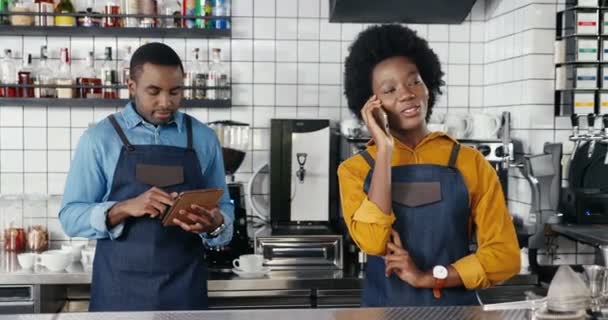 Pretty African American female waitress in apron standing in cafe at counter, talking on mobile phone. Man barrista tapping, texting and scrolling on tablet device on background. Speaking on cellphone - Footage, Video