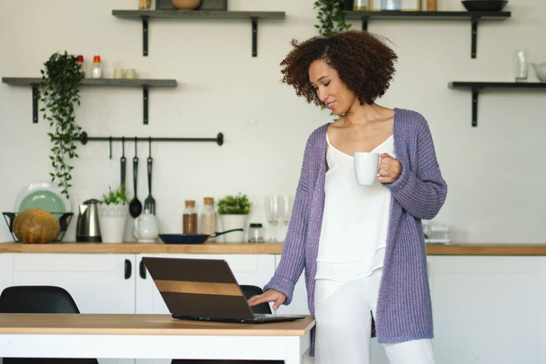 Happy woman holding a cup of coffee and checks mail on his laptop in her kitchen. Young african american female drinking coffee in her kitchen. relaxed morning concept - Fotografie, Obrázek