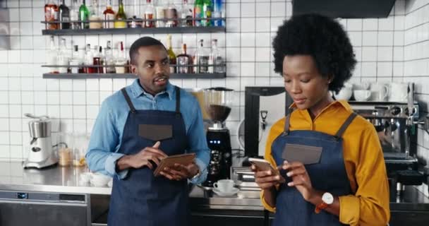Hermosa camarera afroamericana en delantal de pie en la cafetería en el mostrador y mensaje de texto en el teléfono inteligente. Hombre barrista tocando y desplazándose en el dispositivo de la tableta. Sms al teléfono. Chatear en línea. - Metraje, vídeo
