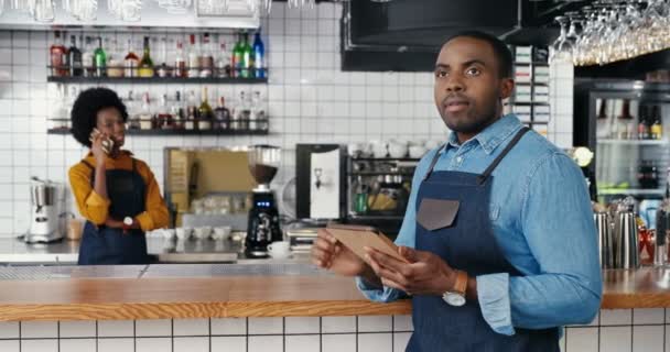 Mužští Afroameričtí barmani používají tabletu v kavárně. Fešák číšník klepe a posouvá se online na gadgetu. Servírka mluví na mobilu v pozadí. Žena mluví na mobilu. - Záběry, video