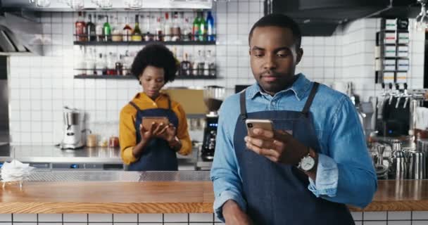 Muž Afroameričtí barmani používají smartphone v kavárně a textovky. Fešák číšník odposlouchává a projíždí se po telefonu. Servírka s tabletovým zařízením na pozadí. Žena držící věcičku. - Záběry, video