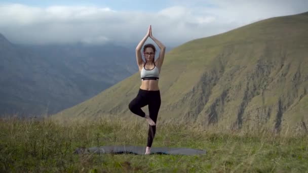 Mujer joven en chándal practica yoga realizando pose de árbol en las montañas. La cámara se mueve creando un efecto de paralaje. - Imágenes, Vídeo