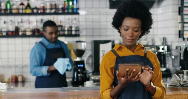 Pretty young African American waitress tapping and scrolling on mobile phone at counter in bar. Woman barista texting message on smartphone. Waiter wiping glasses on background. Cleaning cups in cafe - Footage, Video