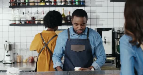 Afro-Amerikalı garson kahve servisi yapıyor ve kafeteryada beyaz bir kadınla konuşuyor. Kahveci adam müşteriyle güzel bir konuşma yapıyor. Erkek barmen bayan müşteriyle sohbet ediyor. Karışık ırklar. - Video, Çekim