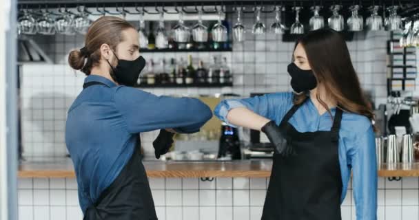 Caucasian young cheerful waiter and waitress in masks, aprons and gloves laughing and touching with elbows like greeting in cafe. Quarantine reopen concept. Male and female baristas meeting in bar. - Footage, Video
