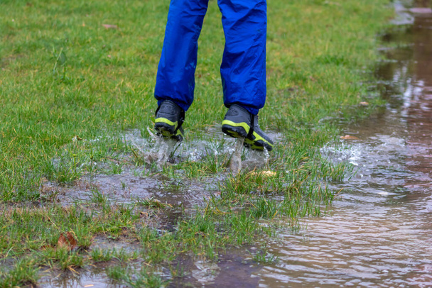 Nogi młodego chłopca w niebieskim wodoodpornym płaszczu przeciwdeszczowym i butach deszczowych skaczą w głębokie kałuże w deszczowy dzień ze szczęściem w rozbryzgu wody jako radosne dzieciństwo i zabawna wycieczka z rodziną - Zdjęcie, obraz