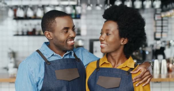 Portret van Afro-Amerikaanse vrolijke barmannen in schorten die elkaar knuffelen en vrolijk naar de camera glimlachen. Gelukkige ober en serveerster in knuffels in café. Vreugdevolle man omarmt vrouw. Barista 's. - Video