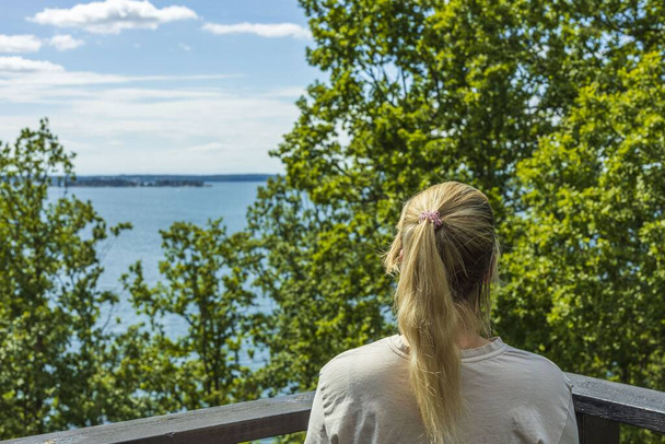 Belle vue estivale sur la mer Baltique paysage. Jeune femme blonde regardant la mer à travers les cimes des arbres verts.  - Photo, image