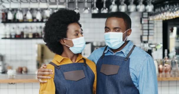 Retrato de un alegre par de camareros afroamericanos con máscaras médicas abrazándose, sonriendo a la cámara felizmente. Feliz camarero y camarera en abrazos en la cafetería. Hombre alegre abrazando mujer baristas - Metraje, vídeo