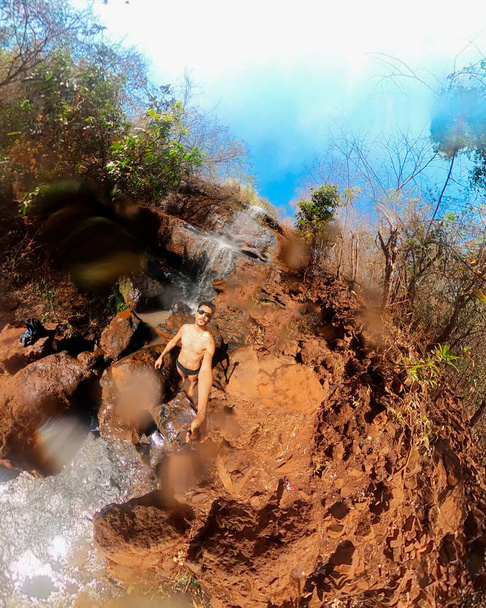 Homme à lunettes et maillots de bain à la cascade avec caméra 360 par une journée ensoleillée - Photo, image