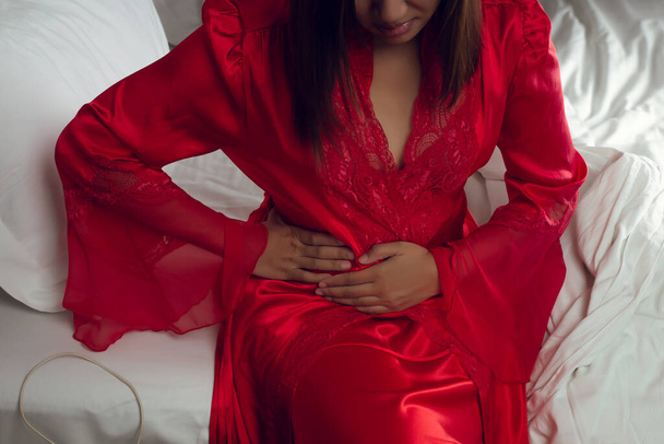 Kobieta w satynowej czerwonej koszuli nocnej cierpiąca na bóle miesiączkowe na łóżku w nocy. Zespół jelita drażliwego lub zespół jelita drażliwego - Zdjęcie, obraz
