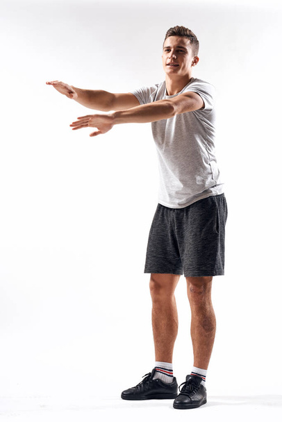A sports man in shorts and a full-length T-shirt does exercises on a light background - Foto, afbeelding