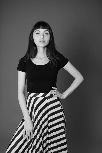 Studio portrait of a pretty brunette woman in a black t-shirt and striped skirt, standing against a plain grey background, looking at camera - Photo, image