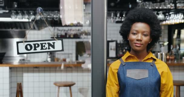 Portrait of beautiful African American young happy woman waitress in apron smiling to camera with table open at cafe entrance outdoor. Pretty cheerful barista standing at bar door with board open. - Footage, Video
