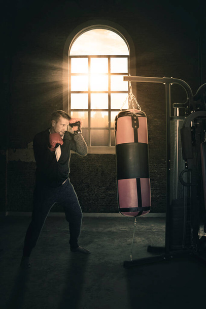Hombre caucásico haciendo ejercicio con una bolsa de boxeo en el centro del gimnasio mientras está de pie con luz solar a través de la ventana - Foto, imagen