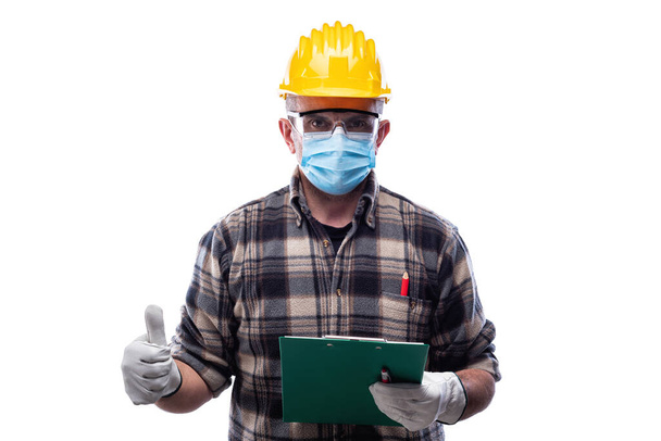 Carpenter worker isolated on white background wears surgical mask to prevent Coronavirus spread, makes OK sign with thumb up. Preventing Pandemic Covid-19 at the workplace. - Photo, image