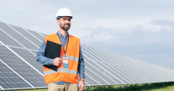 Portrait of male engineer standing on background of solar panels. Caucasian man in special uniform with helmet smiling at camera. - Footage, Video