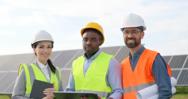 Retrato de ingenieros parados afuera cerca de paneles solares. Tres trabajadores: una mujer y dos hombres con uniforme especial miran a la cámara y sonríen. - Metraje, vídeo