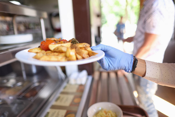 Женщина в синей медицинской перчатке держит белую тарелку с жареным картофелем и тушеными овощами. - Фото, изображение
