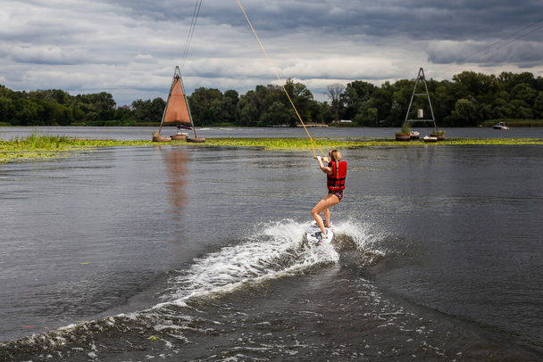 Young pretty slim brunette woman riding wakeboard on in a summer lake. Wakeboarding on cable - Photo, Image