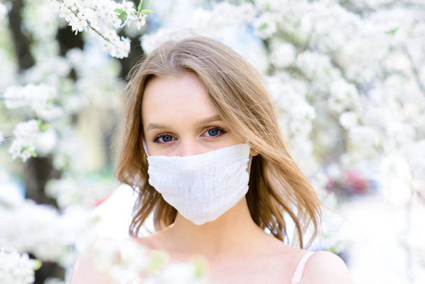 Coronavirus 'un çiçekli maskesiyle çiçek açan kiraz ağacının altında beyaz bir bluz içindeki hassas kızın portresini kapat.. - Fotoğraf, Görsel