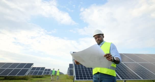 Portret van een jonge Afro-Amerikaanse ingenieur die een zonne-energie plan in zijn handen heeft. Man in speciale uniform en beschermende helm studies papier regeling. - Video