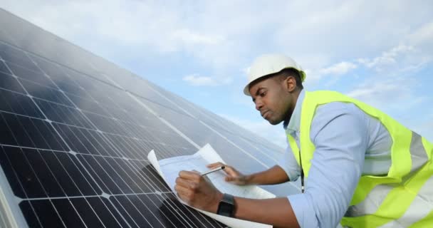 African-American engineer in special uniform making pencil notes on paper diagram leaning on solar panel.Concept of alternative electricity. - Footage, Video
