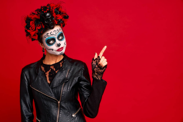 Young woman with sugar skull makeup with a wreath of flowers on her head and skull and black gloves points to a free space. isolated on red background. concept of Halloween or Calavera Catrina. - Photo, Image
