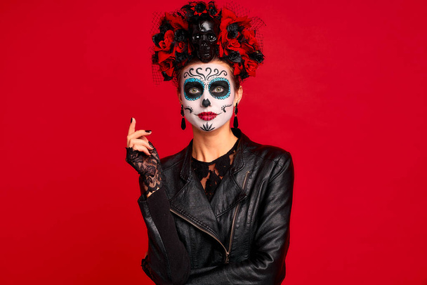 Scary young creepy lady calavera. wears artistic make-up for the feast of all the dead.Wears black leather jacket and lace gloves, dressed as skeleton isolated in red - Fotó, kép