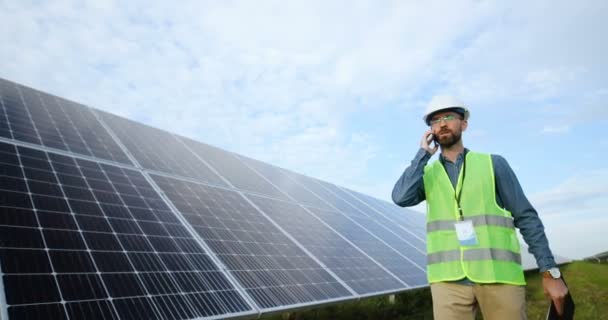 Portrait of Caucasian male engineer talking on phone. Solar farm worker walking along rows with batteries holding documents in his hand. - Footage, Video