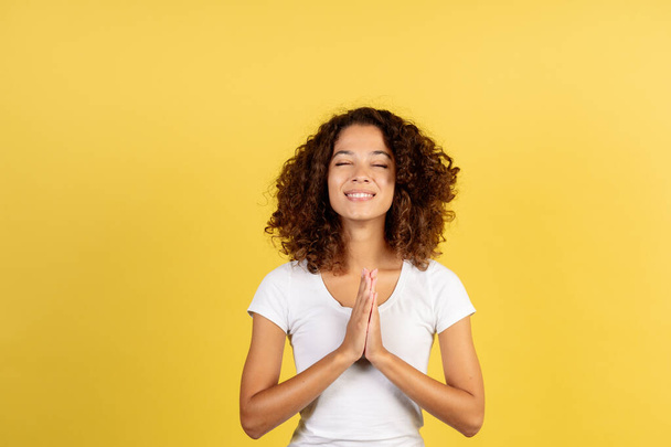 Happy and sincere adult afro american woman make prayer hands, closed eyes and standing on yellow copy space background with nice beaming smile - Photo, Image