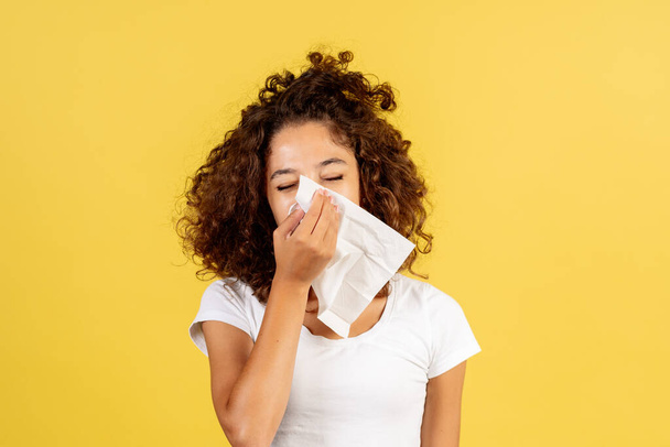 Concept d'allergie saisonnière. Portrait de femme afro-américaine malade adulte avec grippe et nez qui coule tenant une serviette près du visage, debout sur fond d'espace de copie jaune - Photo, image