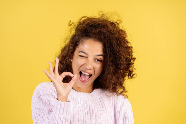 Portrait of happy adult african american woman showing okay sign, wink eye and standing on yellow copy space background in trendy pullover - Foto, Imagen