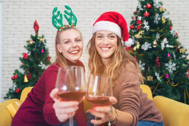 Pareja con copas de vino tinto cerca de la chimenea, hermosa mujer joven caucásica en suéter y con sombrero de santa en la cabeza sentado en el sofá junto al árbol de Navidad - Foto, imagen