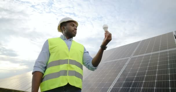 Portret van een Afro-Amerikaanse ingenieur in uniform met een beschermende helm die kijkt naar de gloeilamp die hij in zijn hand heeft. Concept van alternatieve groene energie. - Video