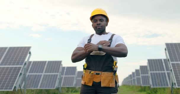 Portrait of young handsome African American craftsman in protective helmet. Man in uniform and with tools standing among solar panels. - Footage, Video