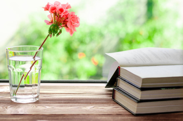 Los libros están listos para leer tumbados en un alféizar de madera junto a un vaso con agua clara en el que hay una flor de geranio rosa - Foto, imagen