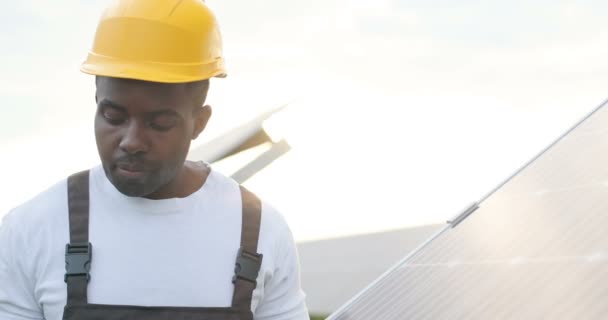 Zbliżenie młodego Afroamerykanina mechanika w żółtym hełmie pracującego na panelu słonecznym. Pojęcie ekologicznej energii elektrycznej. - Materiał filmowy, wideo
