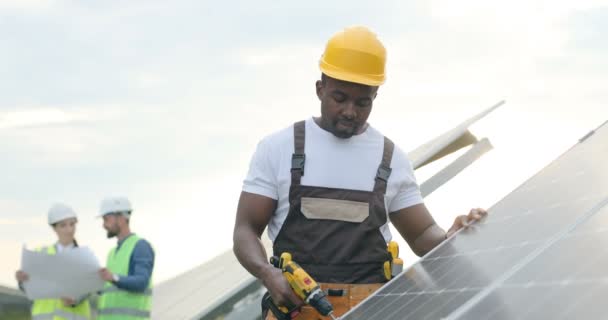 Porträt eines jungen afroamerikanischen Mechanikers mit gelbem Helm, der Löcher in Solarzellen mit Elektrobohrmaschine macht. - Filmmaterial, Video