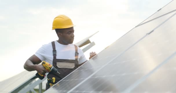 Przystojny młody Afroamerykański mechanik w żółtym hełmie robi dziury w panelu słonecznym z elektrycznym wiertłem. Zwolniony ruch. - Materiał filmowy, wideo