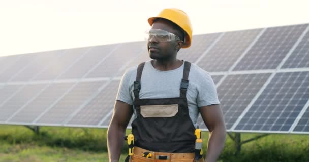 Bel lavoratore afro-americano in piedi nel mezzo di vetro a energia solare e sorrisi. Uomo in casco protettivo con le braccia incrociate pose per macchina fotografica. - Filmati, video
