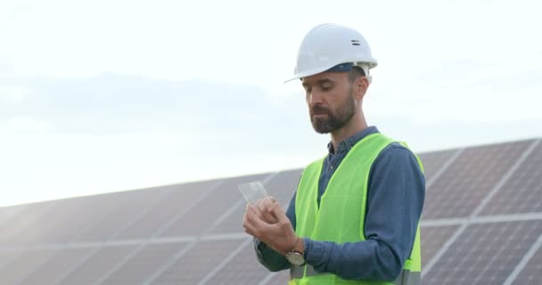 Характерний кавказький чоловічий технік стоїть поза сонячними батареями і оглядає складену табличку. Працює на сонячній сучасній фермі.. - Кадри, відео
