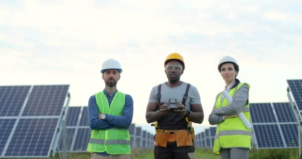 Портрет трех рабочих: женщины и двух мужчин, стоящих на солнечной электростанции. Афроамериканец управляет полетом беспилотника с дистанционным управлением. - Кадры, видео