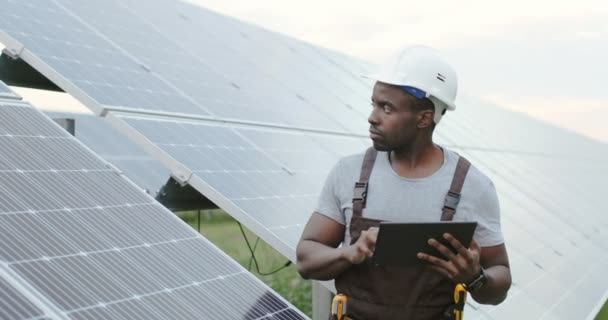 Bellissimo meccanico afroamericano maschio in piedi fuori vicino ai pannelli solari tenendo tablet. Creazione di reti elettriche alternative da parte dell'uomo. - Filmati, video