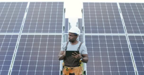 Guapo afroamericano mecánico masculino en uniforme de pie delante de filas de paneles solares sosteniendo tableta digital y mirando hacia otro lado. - Metraje, vídeo
