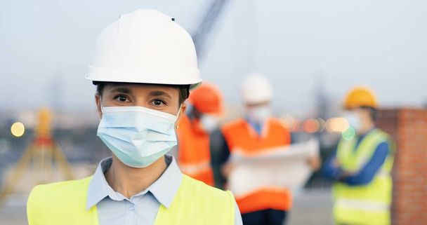 Portrait of Caucasian young beautiful woman constructor in casque and medical mask standing outdoor at construction and looking at camera. Close up of female builder at building in helmet. Pandemic. - Foto, immagini