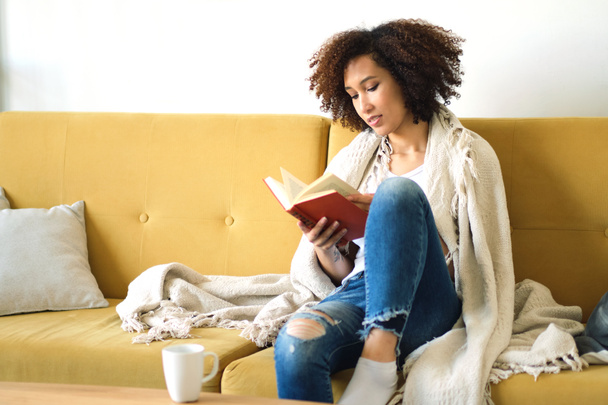 Güzel, Afro-Amerikan bir kadın kitap okuyor. Oturma odasındaki kanepede sırtüstü rahatça yatıyor. Kot pantolonuyla. - Fotoğraf, Görsel